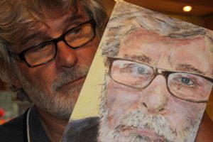 Image of a man with grey hair and glasses holding a painting of himself