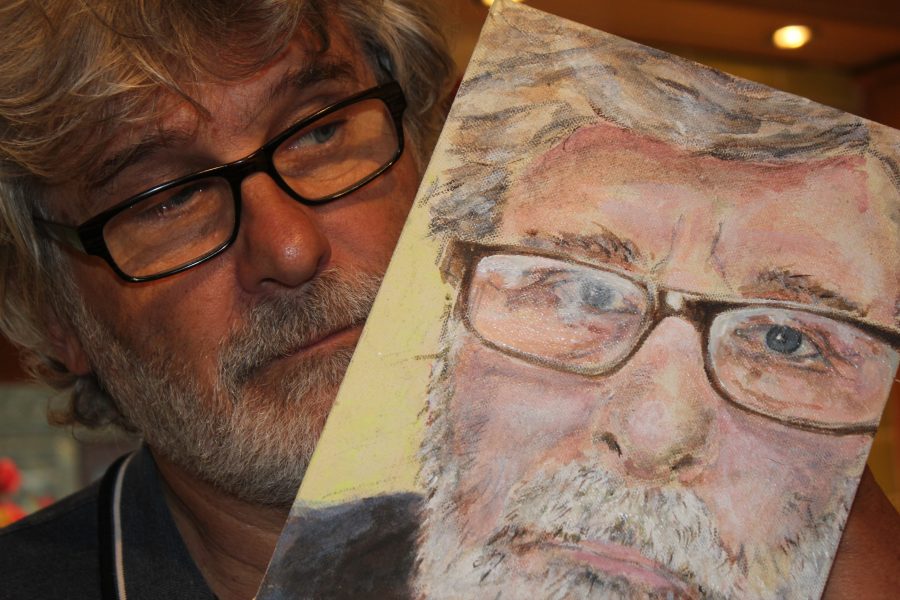 Image of a man with grey hair and glasses holding a painting of himself