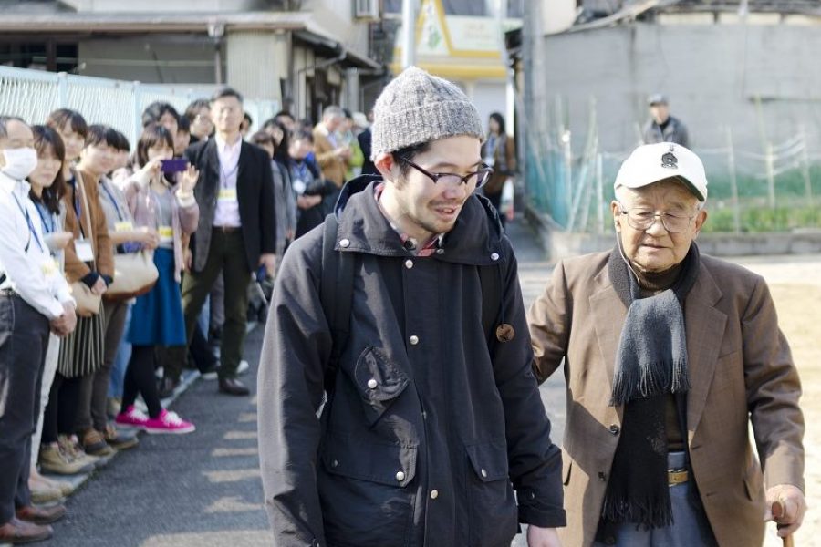 one older and one younger japanese men walking along a path with people gathered in a line watching them.