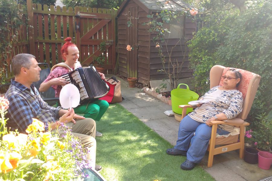 Two men and a woman sitting in a garden. One of the man is playing the accordion.