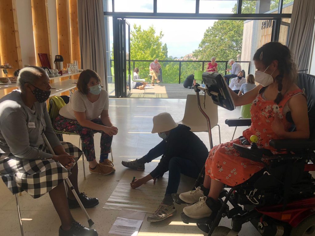 A black older woman and a white woman sit on chairs looking at the woman in front of them on a wheelchair using a text to speech keyboard. Another woman sits on the floor taking notes on a piece of paper. They're all wearing masks.