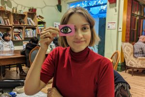 A white woman with short light hair wearing a red jumper and holding a drawing of an eye in front of her eye