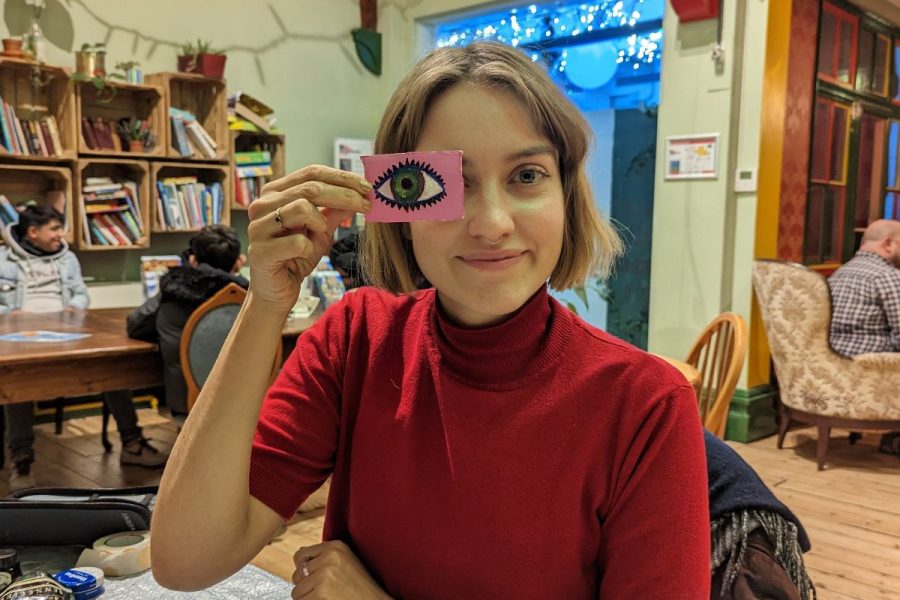 A white woman with short light hair wearing a red jumper and holding a drawing of an eye in front of her eye