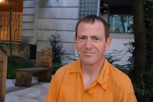 A white man with short dark hair in an orange shirt smiling at the camera