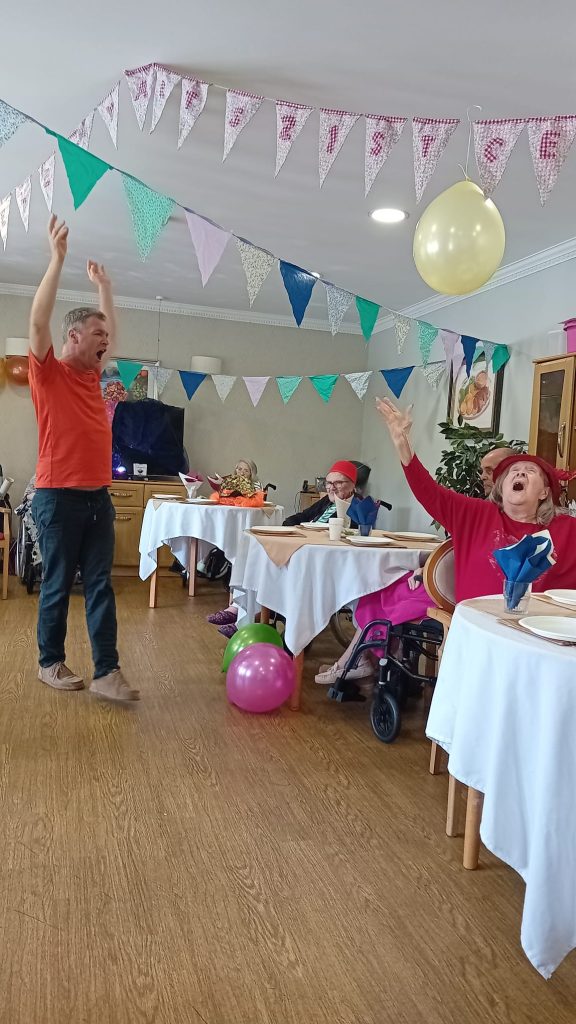 There is bunting on the ceiling. A man in an orange t-shirt raises his arms in the air. An older woman wearing a red bowler hat raises her arms in the air. They are singing.